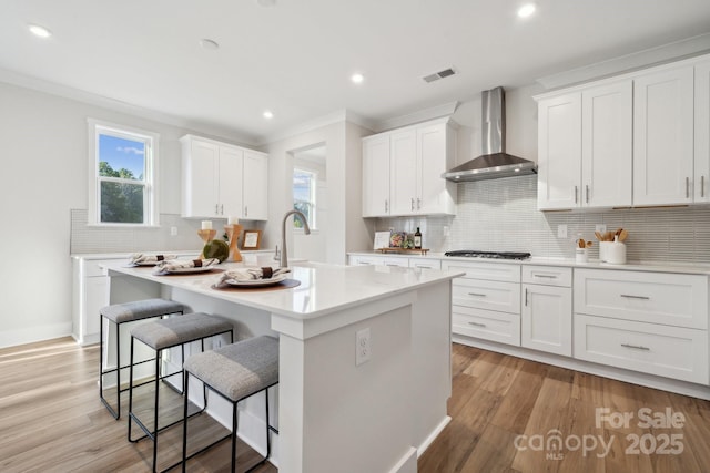 kitchen featuring light hardwood / wood-style floors, wall chimney range hood, white cabinets, and an island with sink