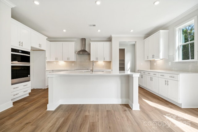 kitchen with white cabinets, wall chimney exhaust hood, ornamental molding, a kitchen island with sink, and light hardwood / wood-style flooring