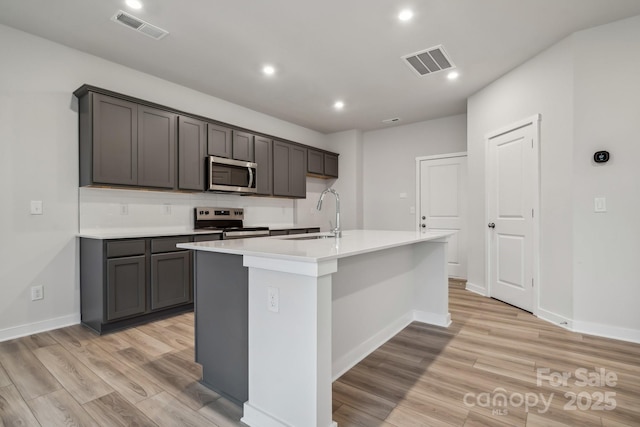 kitchen featuring sink, light hardwood / wood-style floors, gray cabinets, a kitchen island with sink, and appliances with stainless steel finishes