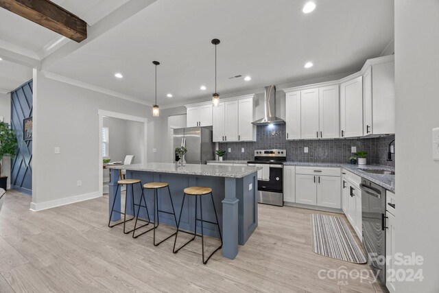 kitchen with appliances with stainless steel finishes, white cabinets, wall chimney exhaust hood, a kitchen island, and decorative light fixtures