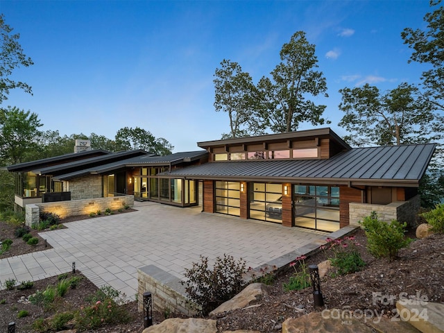 rear view of house featuring a patio area, stone siding, an attached garage, and a standing seam roof