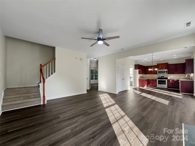 unfurnished living room with ceiling fan with notable chandelier, dark wood-type flooring, visible vents, baseboards, and stairway