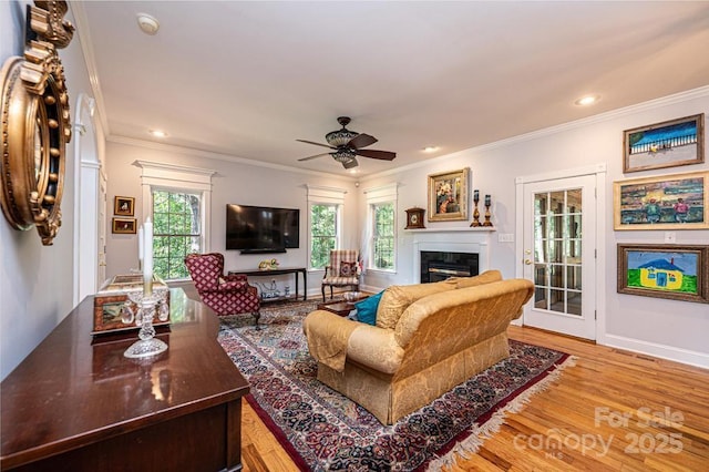 living area featuring baseboards, a glass covered fireplace, ceiling fan, wood finished floors, and crown molding