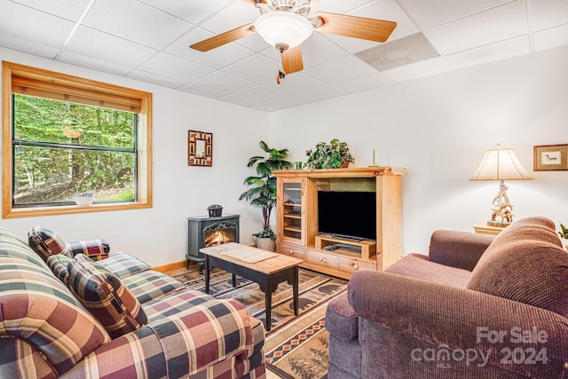 living area featuring a ceiling fan, wood finished floors, a paneled ceiling, baseboards, and a wood stove