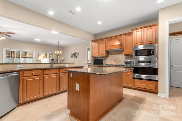 kitchen featuring dark stone countertops, custom exhaust hood, ceiling fan with notable chandelier, stainless steel appliances, and a kitchen island