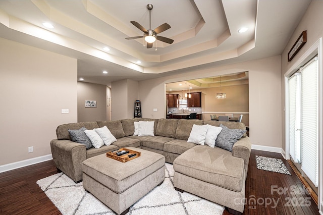 living room with ceiling fan with notable chandelier, dark wood-type flooring, and a tray ceiling