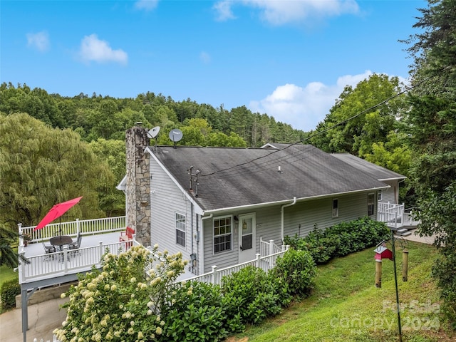 back of house with a wooded view, a yard, roof with shingles, a chimney, and a deck