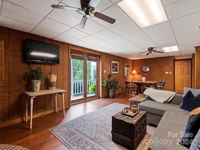 living room with a paneled ceiling, wood finished floors, wood walls, and a ceiling fan