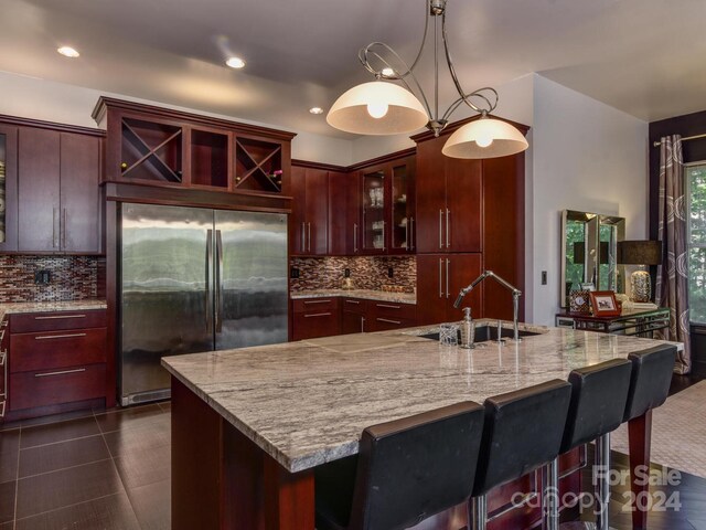 kitchen featuring decorative light fixtures, a center island with sink, backsplash, stainless steel refrigerator, and light stone counters