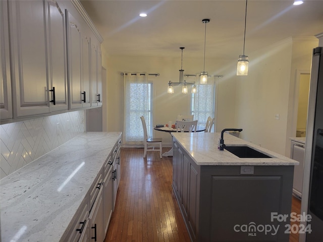 kitchen with a kitchen island with sink, dark wood-type flooring, sink, light stone countertops, and pendant lighting