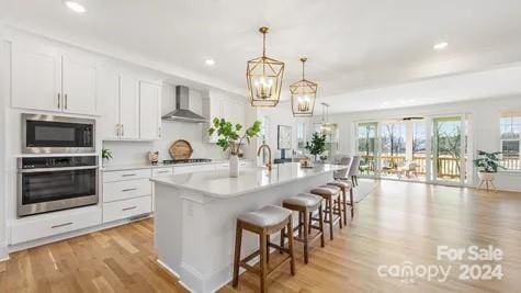 kitchen with white cabinets, appliances with stainless steel finishes, a kitchen island with sink, wall chimney exhaust hood, and decorative light fixtures