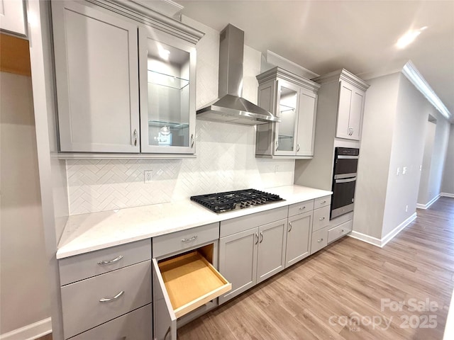 kitchen featuring gray cabinetry, stainless steel gas cooktop, decorative backsplash, light wood-style flooring, and wall chimney exhaust hood