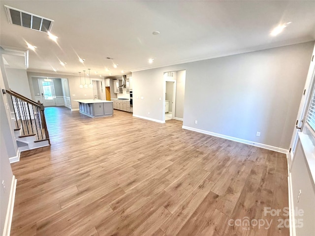 unfurnished living room with light wood-type flooring, visible vents, stairway, crown molding, and baseboards