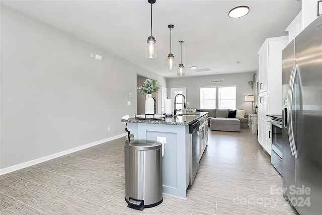 kitchen featuring hanging light fixtures, dark stone countertops, stainless steel appliances, white cabinetry, and a center island with sink