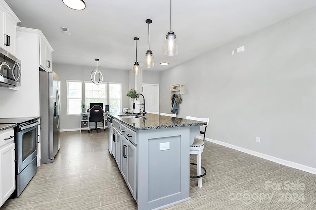 kitchen featuring white cabinets, dark stone countertops, hanging light fixtures, stainless steel appliances, and an island with sink