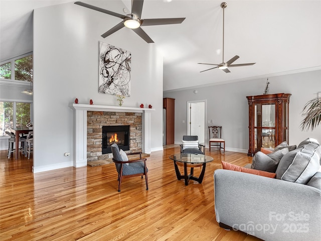 living room with high vaulted ceiling, ceiling fan, a stone fireplace, and light hardwood / wood-style floors