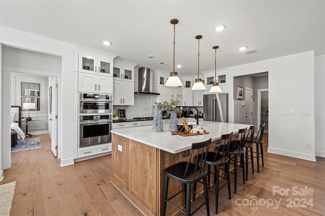 kitchen with white cabinets, decorative light fixtures, a center island with sink, wall chimney range hood, and stainless steel appliances