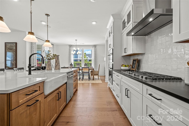 kitchen with white cabinetry, light hardwood / wood-style flooring, stainless steel appliances, wall chimney exhaust hood, and decorative light fixtures