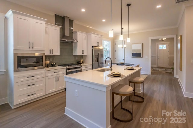 kitchen with sink, wall chimney exhaust hood, stainless steel appliances, an island with sink, and pendant lighting