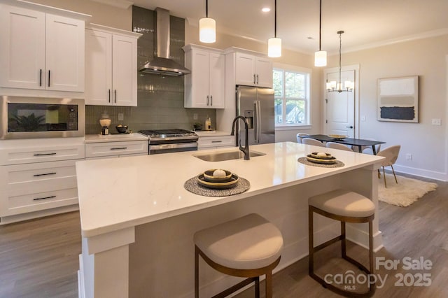 kitchen with hanging light fixtures, wall chimney range hood, stainless steel appliances, and a kitchen island with sink