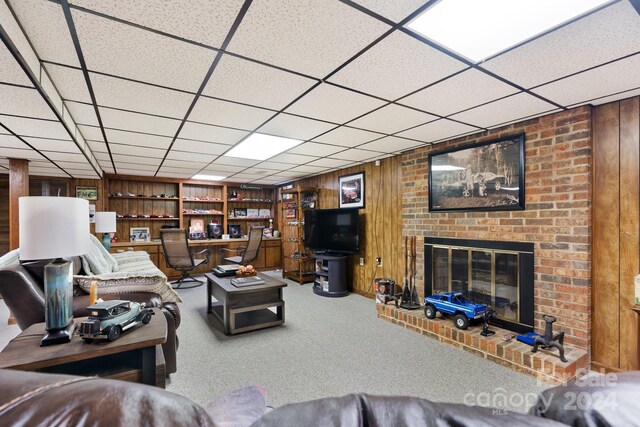 living room with carpet, a brick fireplace, wood walls, and a drop ceiling