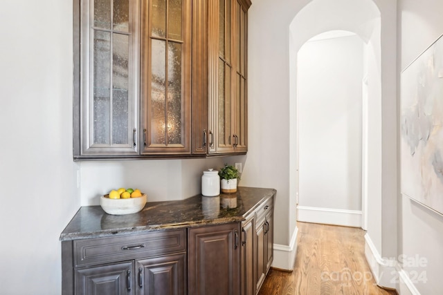 bar with dark stone countertops, dark brown cabinets, and light wood-type flooring