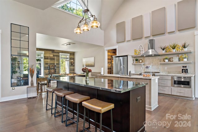 kitchen with dark hardwood / wood-style flooring, appliances with stainless steel finishes, a large island with sink, high vaulted ceiling, and wall chimney exhaust hood