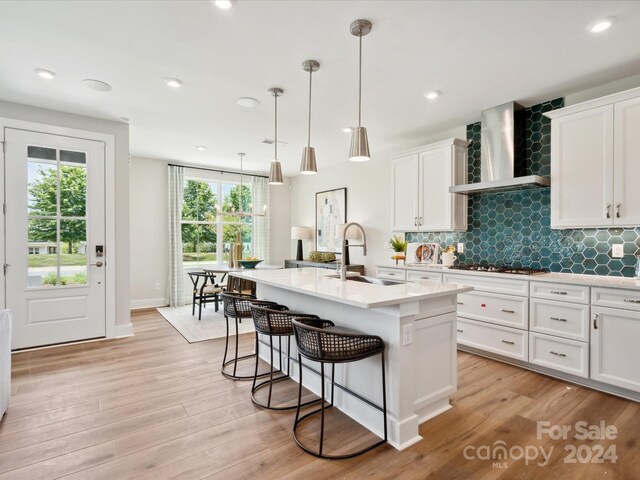 kitchen with light hardwood / wood-style floors, an island with sink, hanging light fixtures, wall chimney range hood, and white cabinets