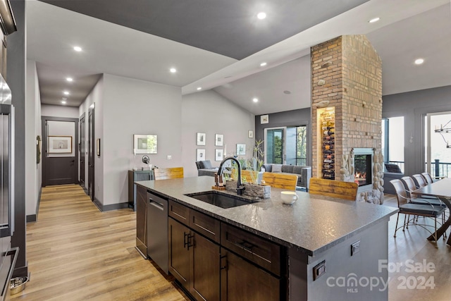 kitchen with a stone fireplace, a kitchen island with sink, sink, lofted ceiling, and dark stone countertops