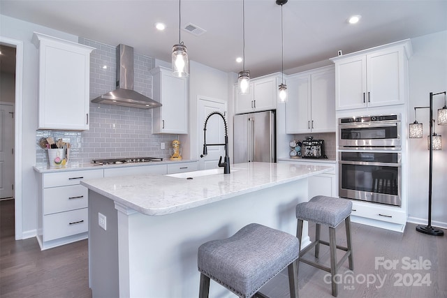 kitchen with appliances with stainless steel finishes, a kitchen island with sink, wall chimney exhaust hood, and white cabinetry