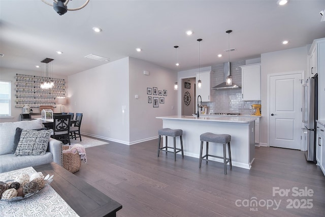 kitchen with hanging light fixtures, stainless steel appliances, white cabinets, a center island with sink, and wall chimney exhaust hood