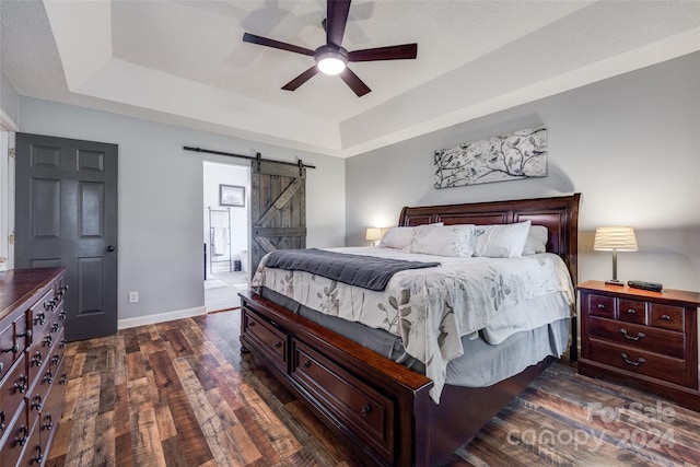 bedroom featuring connected bathroom, dark hardwood / wood-style flooring, ceiling fan, a tray ceiling, and a barn door