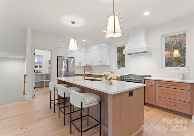 kitchen with stainless steel appliances, premium range hood, an island with sink, light hardwood / wood-style floors, and white cabinets