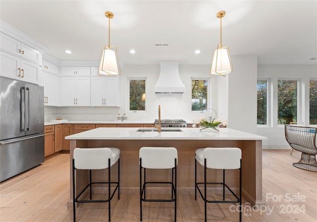 kitchen featuring white cabinets, custom range hood, stainless steel refrigerator, and an island with sink