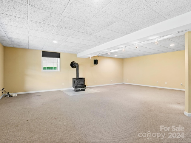 basement with rail lighting, carpet flooring, a paneled ceiling, and a wood stove