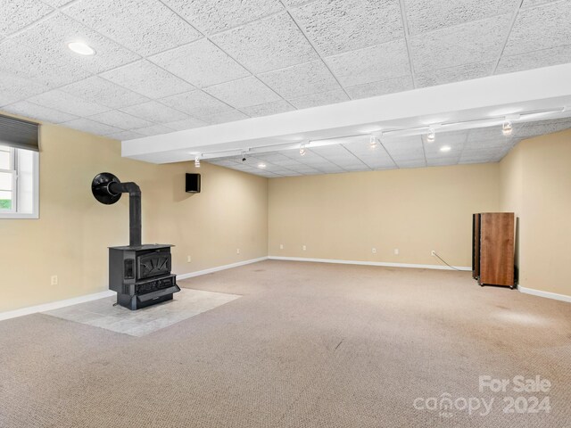 basement featuring a paneled ceiling, a wood stove, and carpet floors