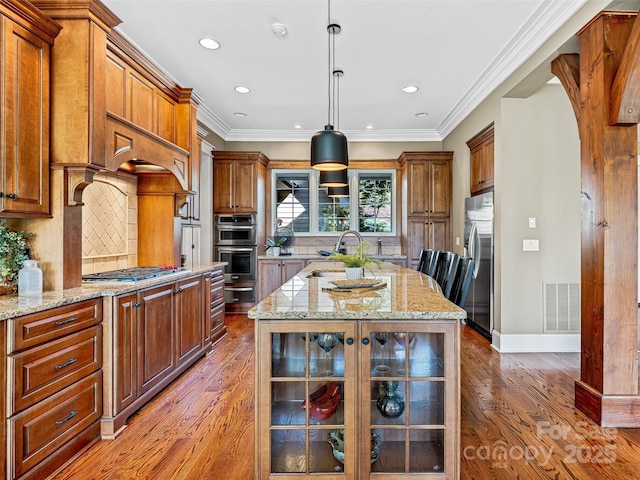 kitchen with visible vents, glass insert cabinets, appliances with stainless steel finishes, light stone countertops, and a kitchen island with sink