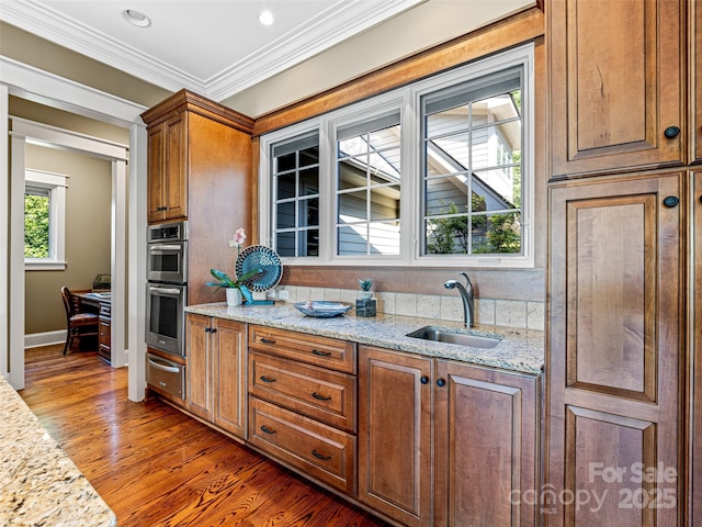 kitchen with stainless steel double oven, a sink, light stone countertops, dark wood-style floors, and crown molding