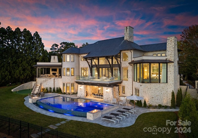 back house at dusk featuring a balcony, a lawn, a patio area, and a swimming pool with hot tub