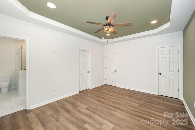 unfurnished bedroom with a tray ceiling, ensuite bathroom, ceiling fan, and hardwood / wood-style flooring