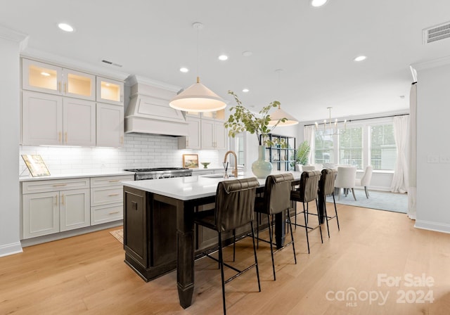 kitchen with custom exhaust hood, hanging light fixtures, light hardwood / wood-style floors, and an island with sink