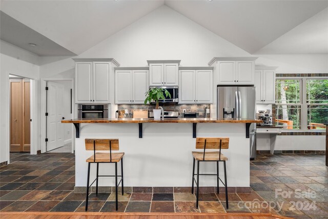 kitchen featuring high vaulted ceiling, a breakfast bar, stainless steel appliances, an island with sink, and stone tile floors