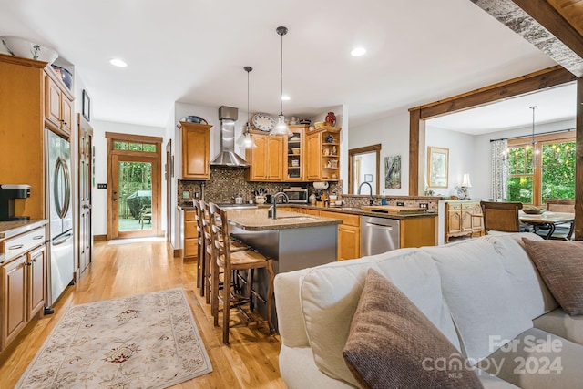 kitchen featuring decorative light fixtures, a kitchen island with sink, light hardwood / wood-style flooring, wall chimney range hood, and appliances with stainless steel finishes