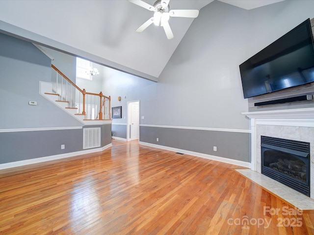 unfurnished living room featuring a tiled fireplace, ceiling fan with notable chandelier, high vaulted ceiling, and light hardwood / wood-style flooring