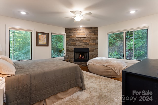 bedroom featuring wood-type flooring, a stone fireplace, multiple windows, and ceiling fan