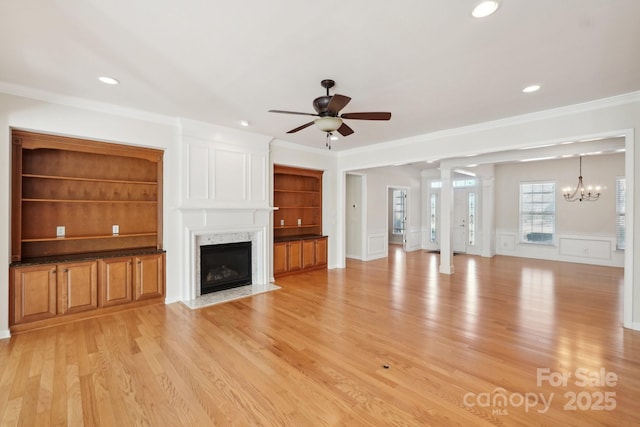 unfurnished living room with a premium fireplace, light wood-type flooring, crown molding, ceiling fan with notable chandelier, and built in features