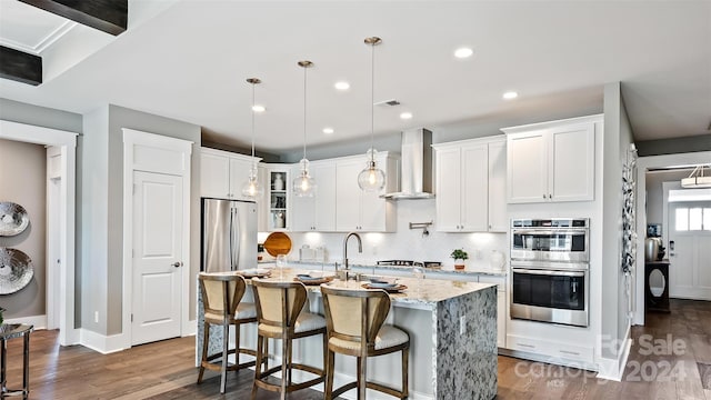 kitchen with wall chimney range hood, stainless steel appliances, dark hardwood / wood-style flooring, and a kitchen island with sink