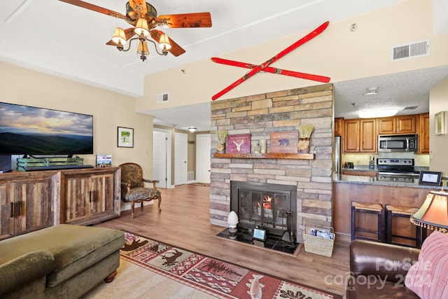 living room featuring a textured ceiling, ceiling fan, a stone fireplace, and light wood-type flooring