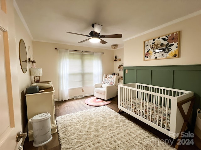 bedroom with crown molding, dark wood-type flooring, a nursery area, and ceiling fan