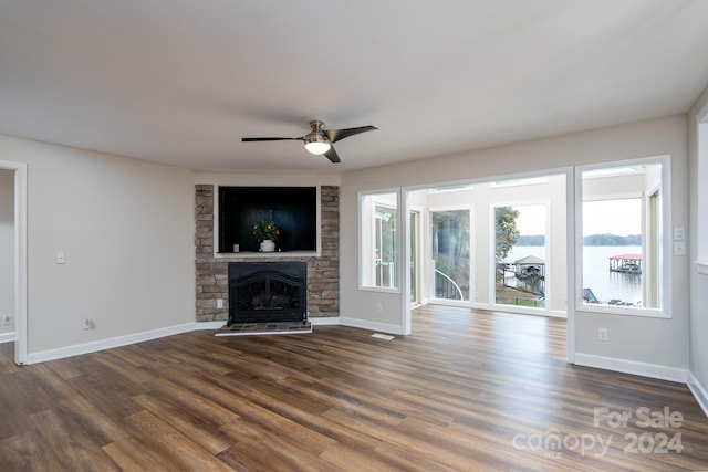 unfurnished living room with dark hardwood / wood-style flooring, a water view, a stone fireplace, and ceiling fan